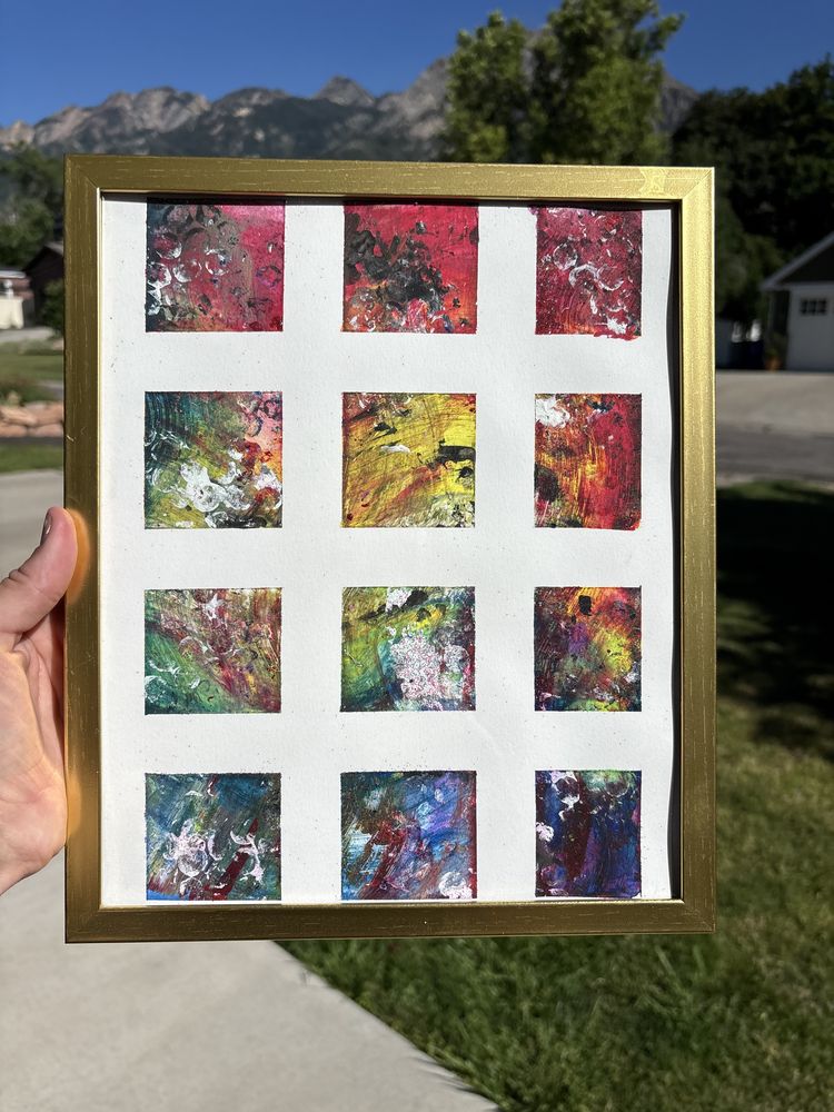 A gold frame held up outdoors with mountains behind it, holding a grid of twelve small abstract paintings in reds, greens and blues
