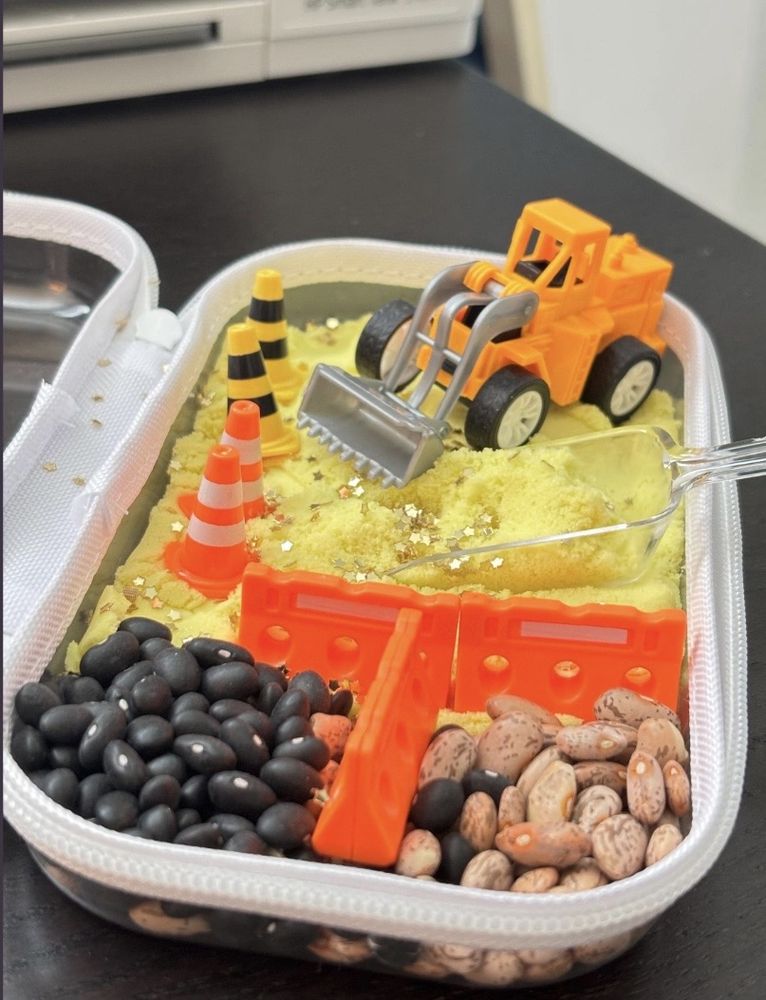 An open Dig In! kit in use: yellow kinetic sand with gold stars, a toy digger, traffic cones, orange barriers and sections of black and pinto beans
