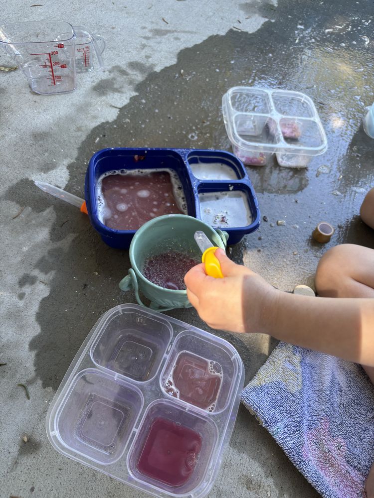 A child's hands scooping a purple potion mixture into a small bucket outdoors, with divided containers of colored liquids nearby