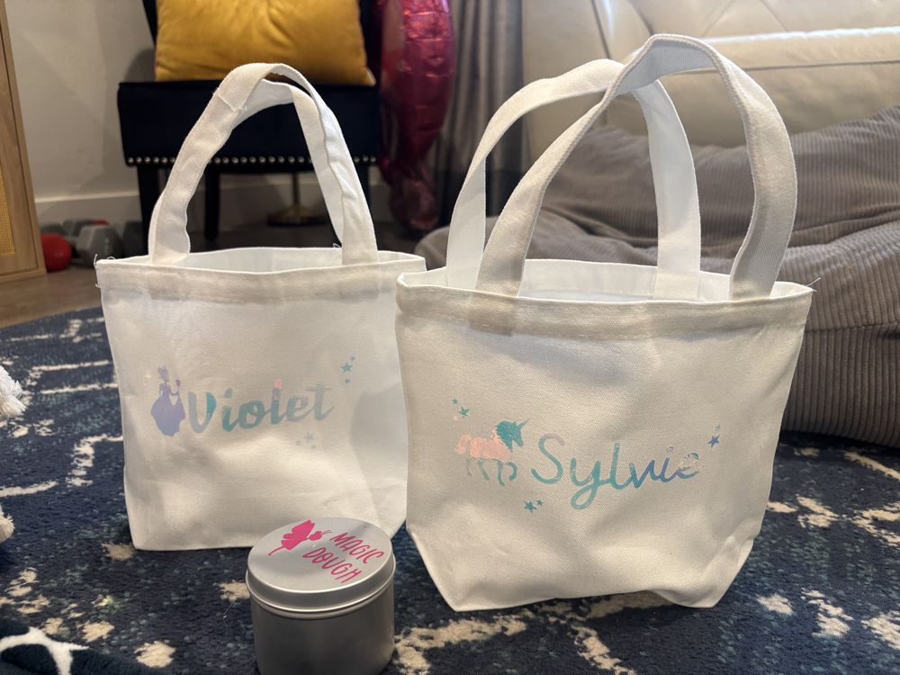 Two small white canvas tote bags personalized with the names Violet and Sylvie in pastel lettering, beside a tin of magic dough